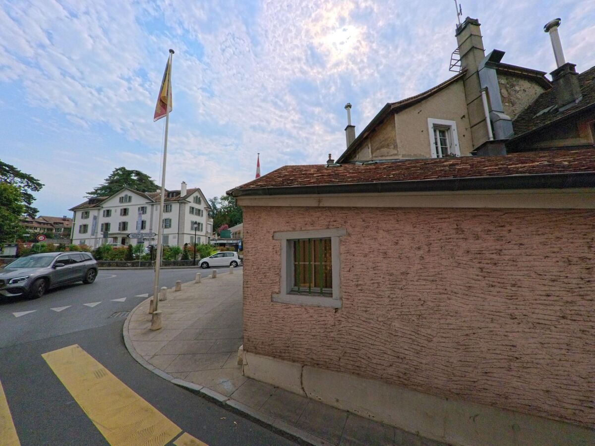 Eine Strasse in Cologny (1223, Kanton Genf) verläuft zwischen historischen Gebäuden, Fahnenmasten und einem weissen Wohnhaus im Hintergrund.