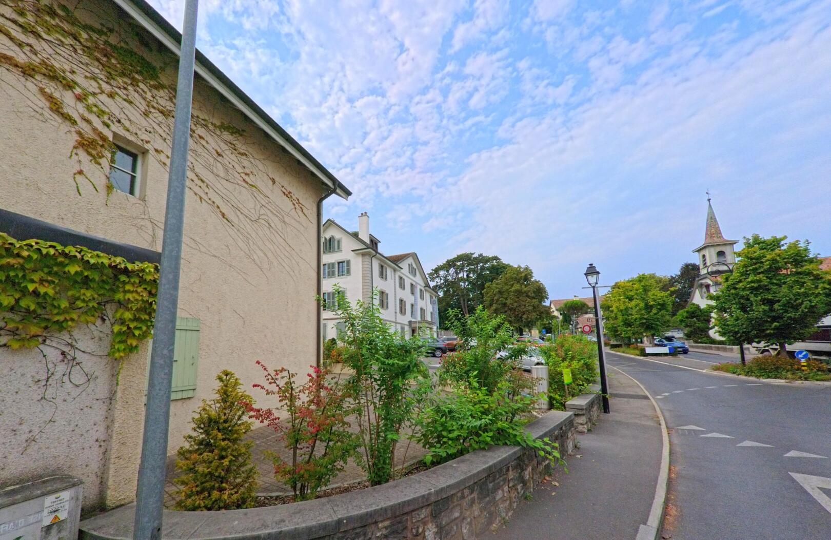 Eine Strasse in Cologny (1223, Kanton Genf) führt an bepflanzten Mauern vorbei zu einem Platz mit Kirche und weissem Wohnhaus.