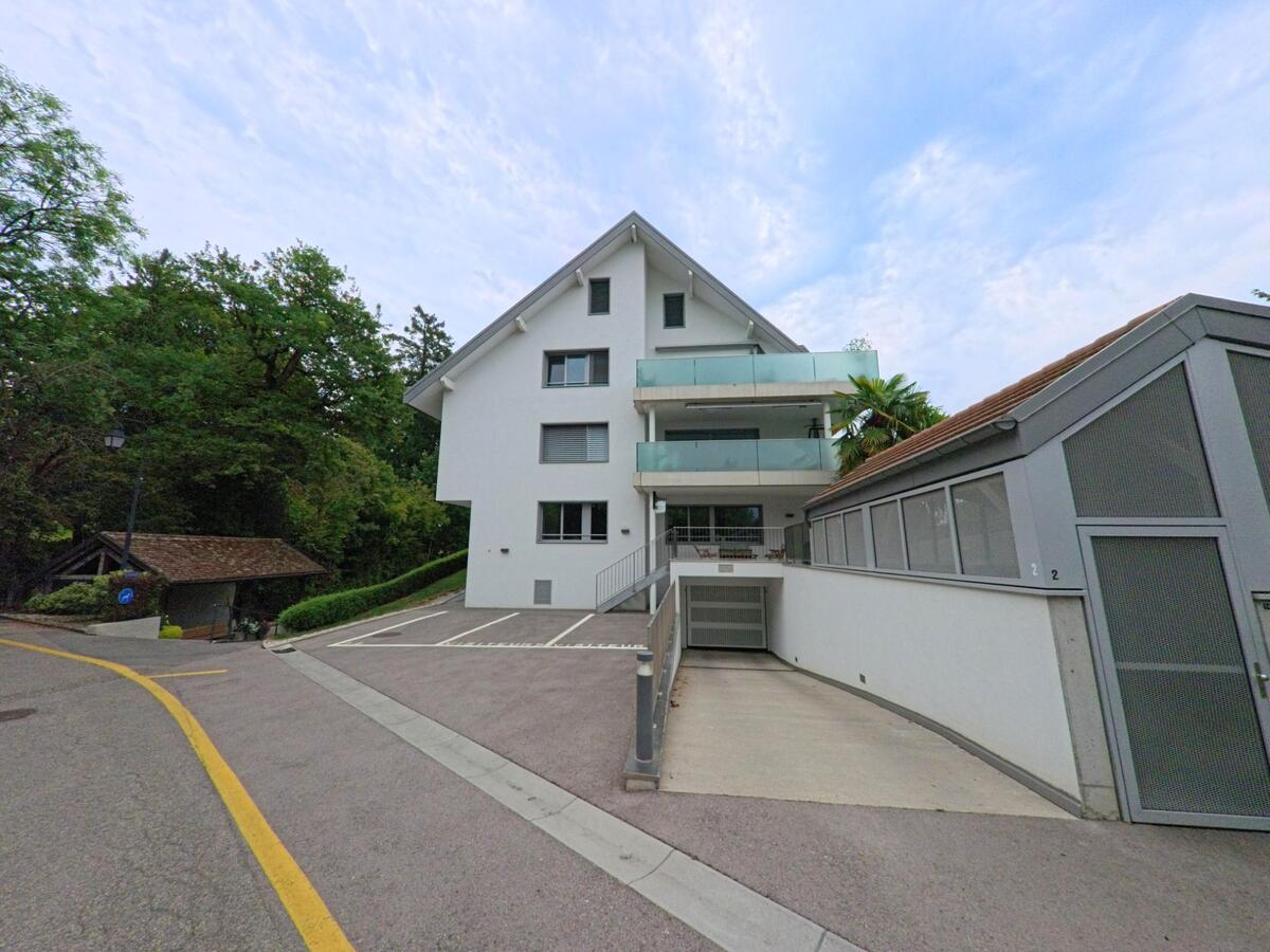 In Commugny (1291, Kanton Waadt) steht ein modernes weisses Mehrfamilienhaus mit Balkonen und Tiefgaragenzufahrt am Strassenrand.