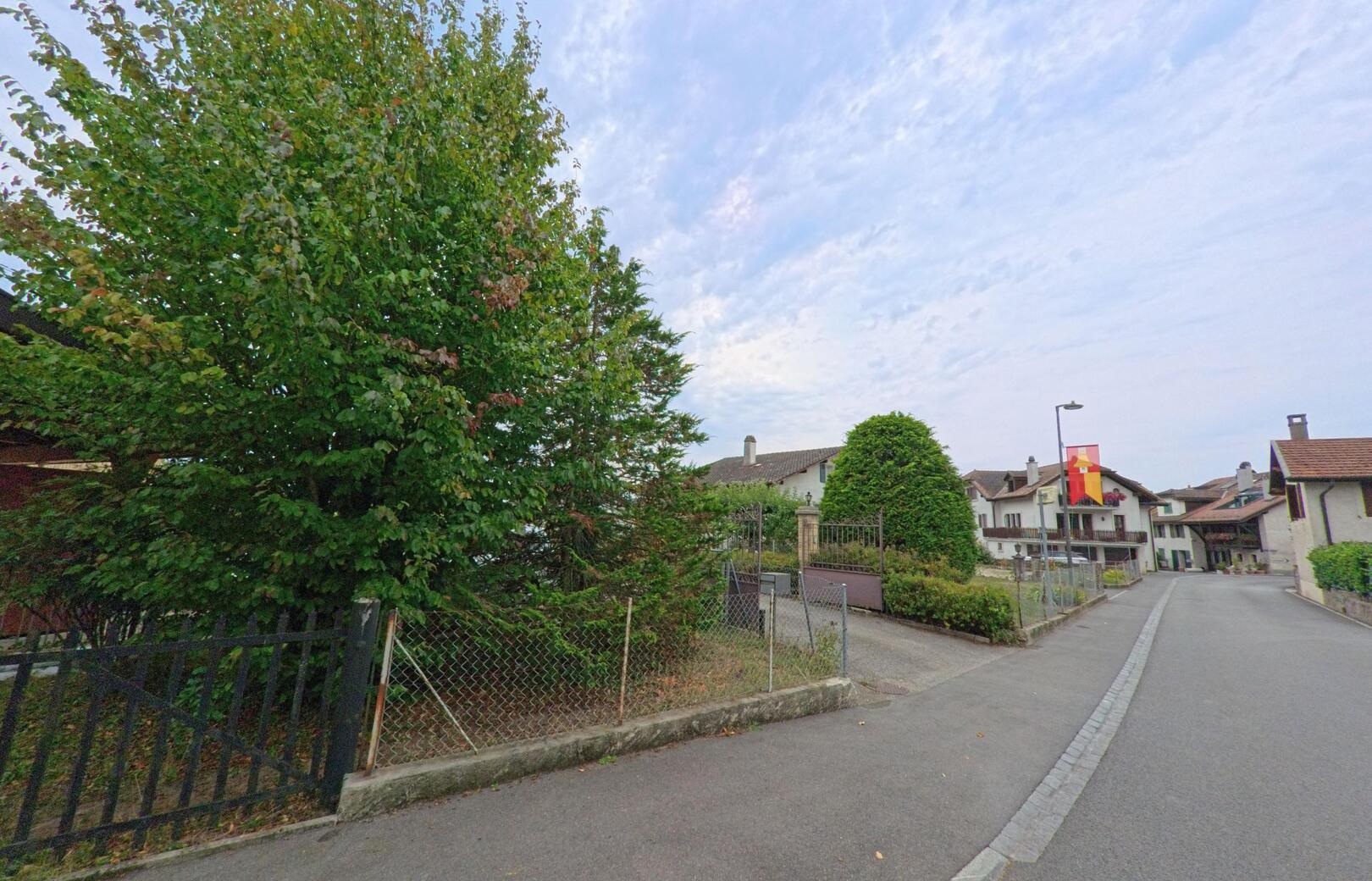 Eine ruhige Seitenstrasse in Commugny (1291, Kanton Waadt) fuehrt an traditionellen Wohnhaeusern mit Ziegeldach und gepflegten Gaerten vorbei.