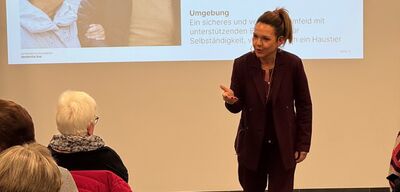 Vor einem projizierten Vortrag spricht eine Referentin mit dem Publikum in Zollikon (8702, Kanton Zürich) und erklaert InhVor einem projizierten Vortrag spricht eine Referentin mit dem Publikum in Zollikon (8702, Kanton Zürich) und erklaert Inhalte zu einem fachlichen Thema.alte zu einem fachlichen Thema.