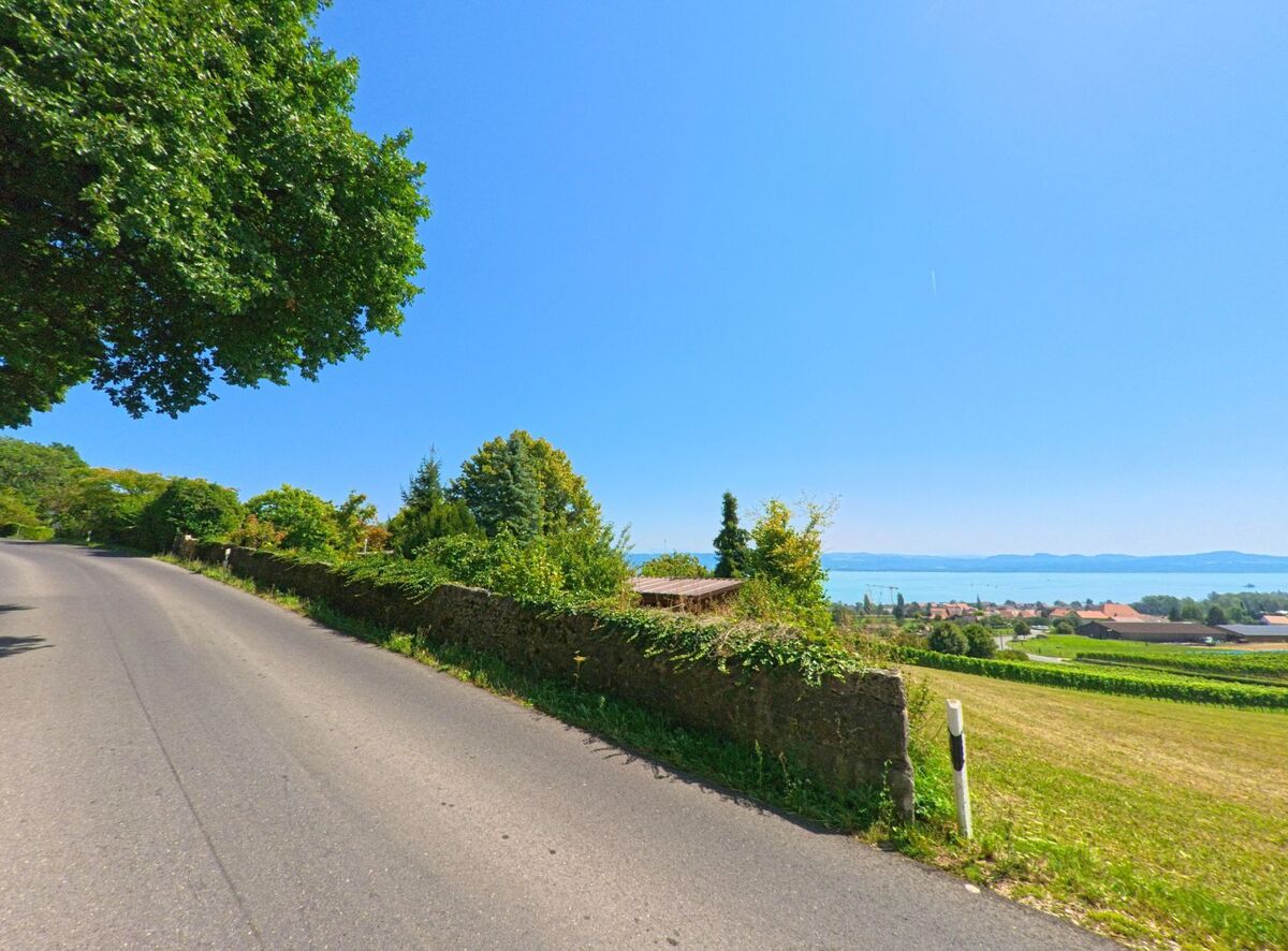 Eine ansteigende Landstrasse in Concise (1426, Kanton Waadt) fuehrt vorbei an einer Steinmauer mit Blick auf den Neuenburgersee und die umliegenden Felder.