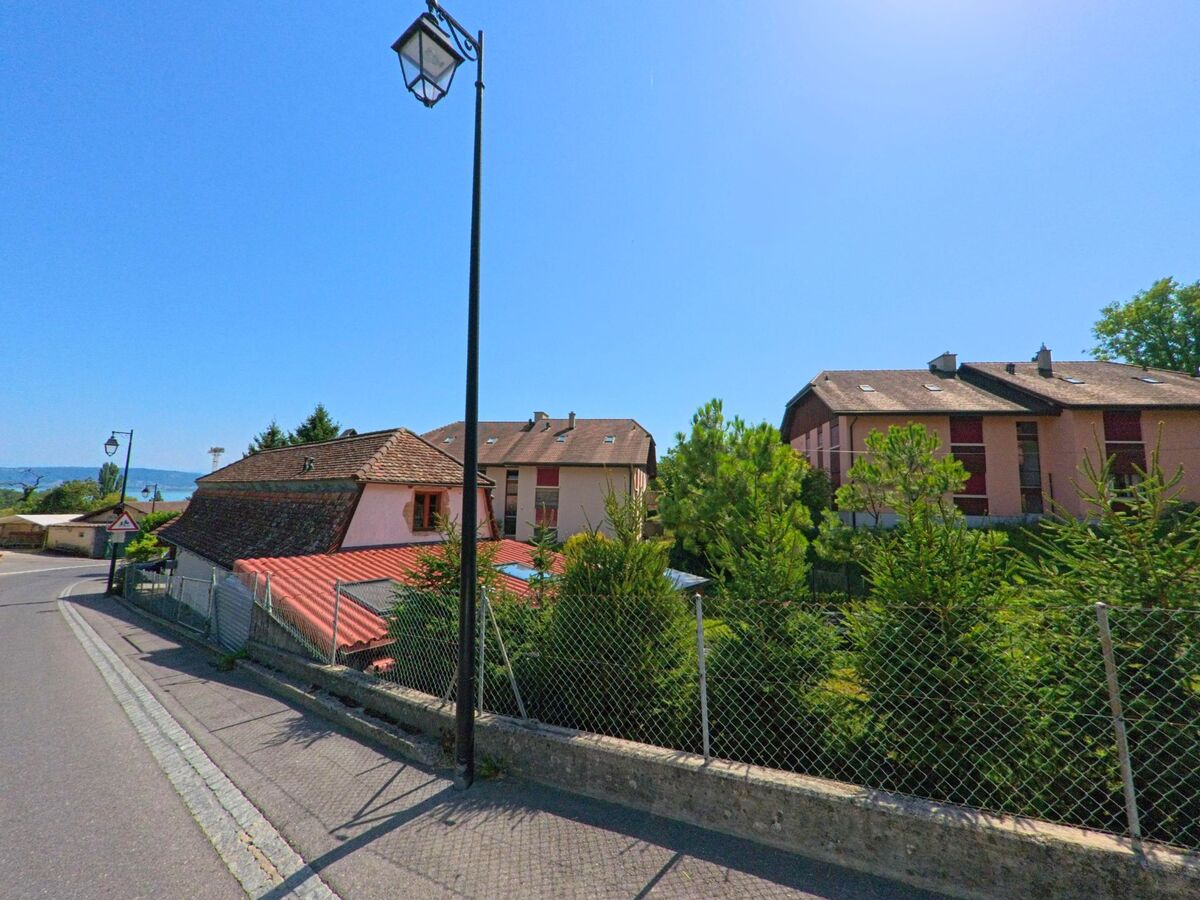 Mehrfamilienhaeuser mit roten Fassaden und ein traditionelles Gebaeude mit Schindeldach stehen in Concise (1426, Kanton Waadt) an einer abfallenden Strasse mit Blick auf den See.