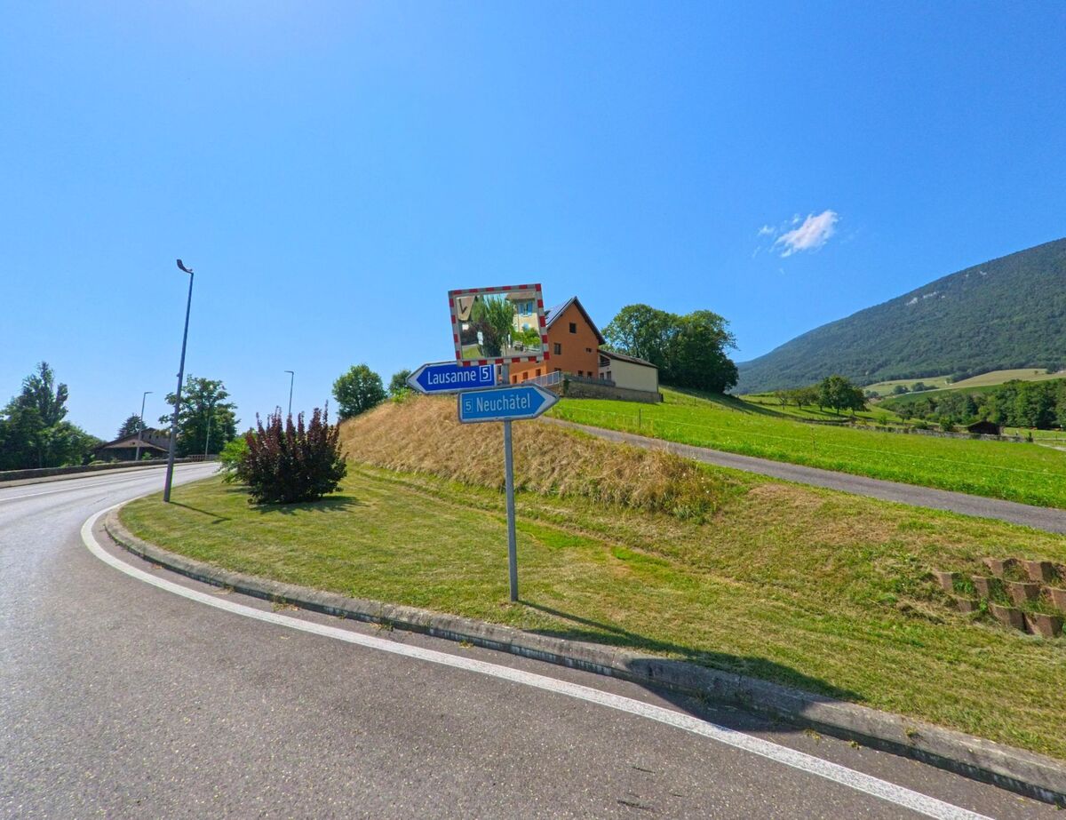 Ein Wegweiser an der Ausfahrt von Concise (1426, Kanton Waadt) zeigt die Richtungen nach Lausanne und Neuchatel, im Hintergrund sind Haeuser und gruene Huegel zu sehen.