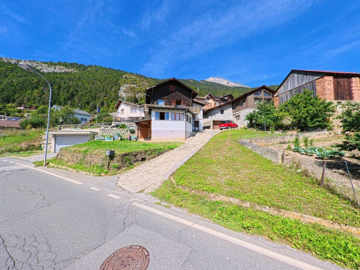 Ein traditionelles Wohnhaus mit Holzbalkon und angrenzenden Gaerten liegt an einem Hang in Conthey (1976, Kanton Wallis).