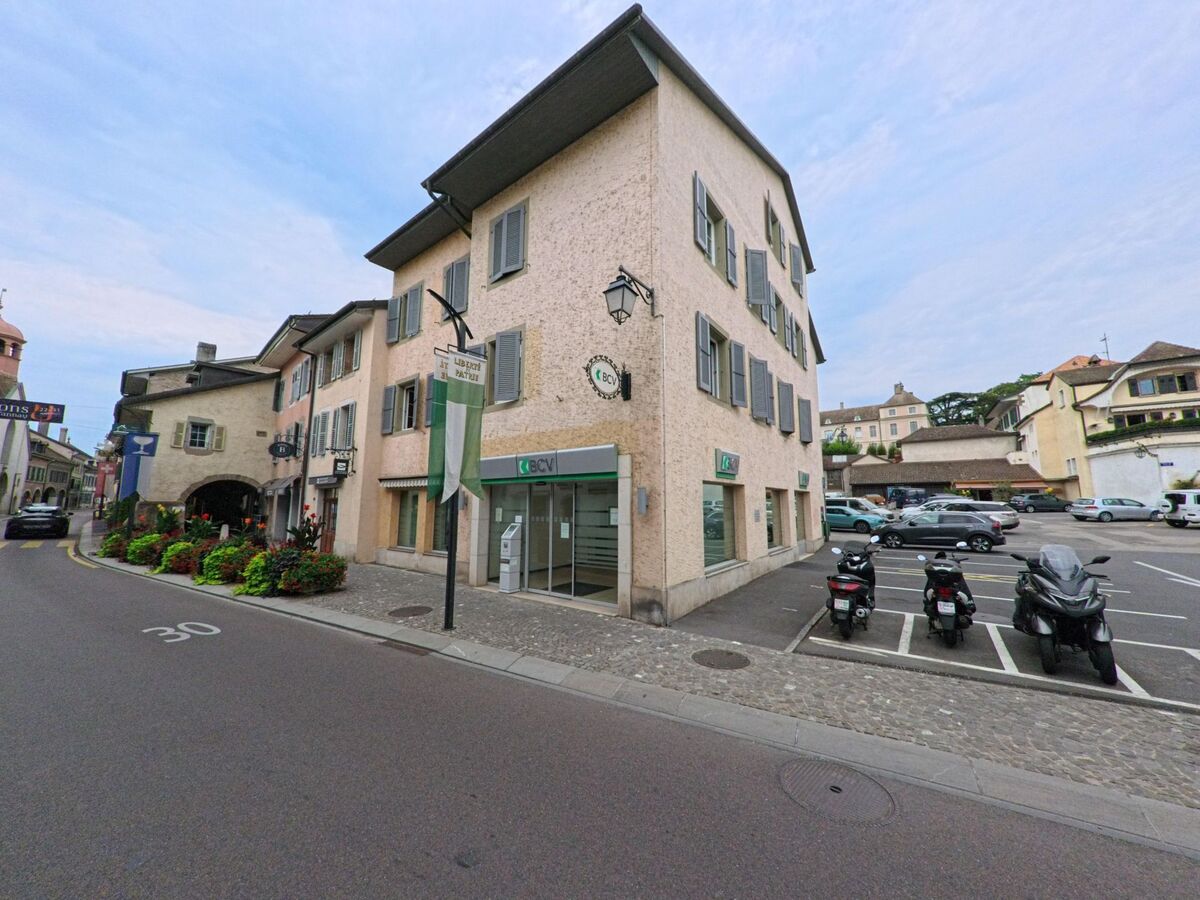 In Coppet (1296, Kanton Waadt) liegt an einer gepflasterten Strasse ein Gebaeude mit Uhr und Bankfiliale, daneben ein kleiner Parkplatz mit Motorraedern.