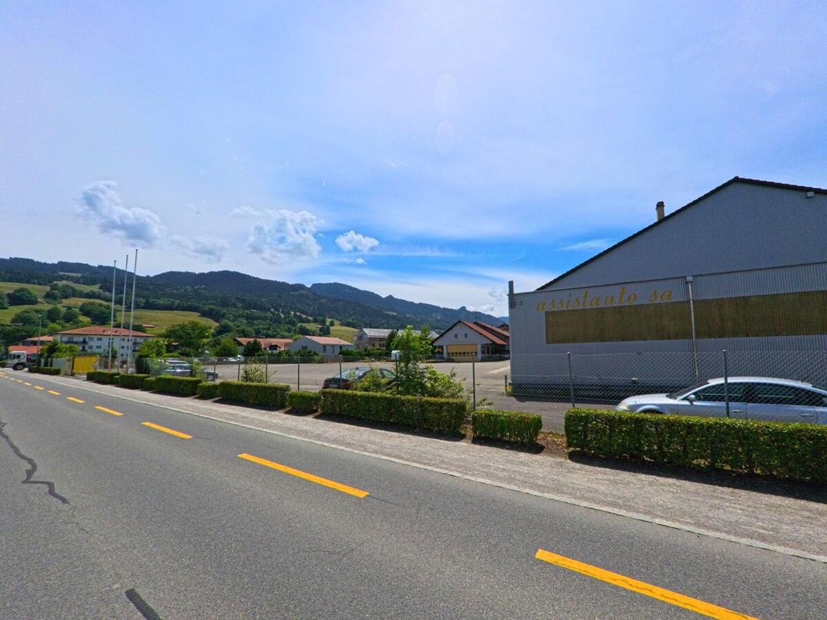 In Corbières (PLZ 1647, Kanton Freiburg) liegt eine Werkhalle mit dem Schriftzug „assistauto sa“ an einer breiten Strasse, im Hintergrund erstreckt sich die Hügellandschaft des Freiburger Voralpengebiets.