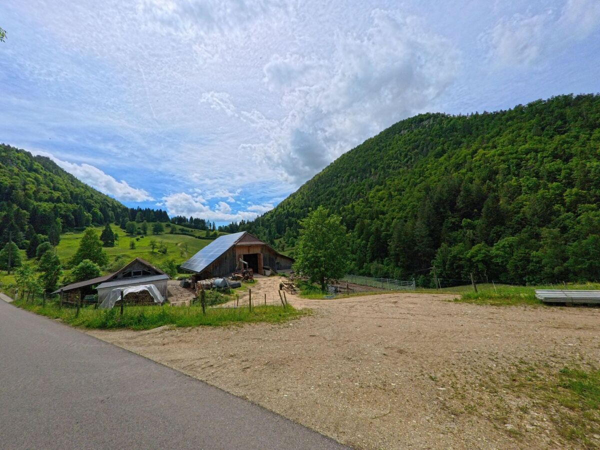 Zwischen zwei bewaldeten Hügeln in Corcelles (2747, Kanton Bern) liegt ein Bauernhof mit Scheune und Solardach, eingerahmt von Wiesen und einer schmalen Strasse im Vordergrund.