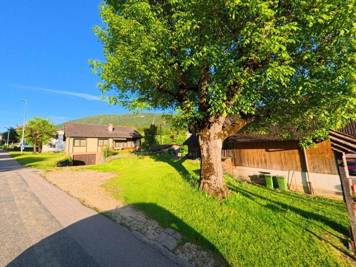 In Corcelles (2747, Kanton Bern) steht ein grosser Laubbaum vor einem kleinen Wohnhaus und einer Holzscheune, während das warme Sonnenlicht die Wiese und die Hügel im Hintergrund beleuchtet.