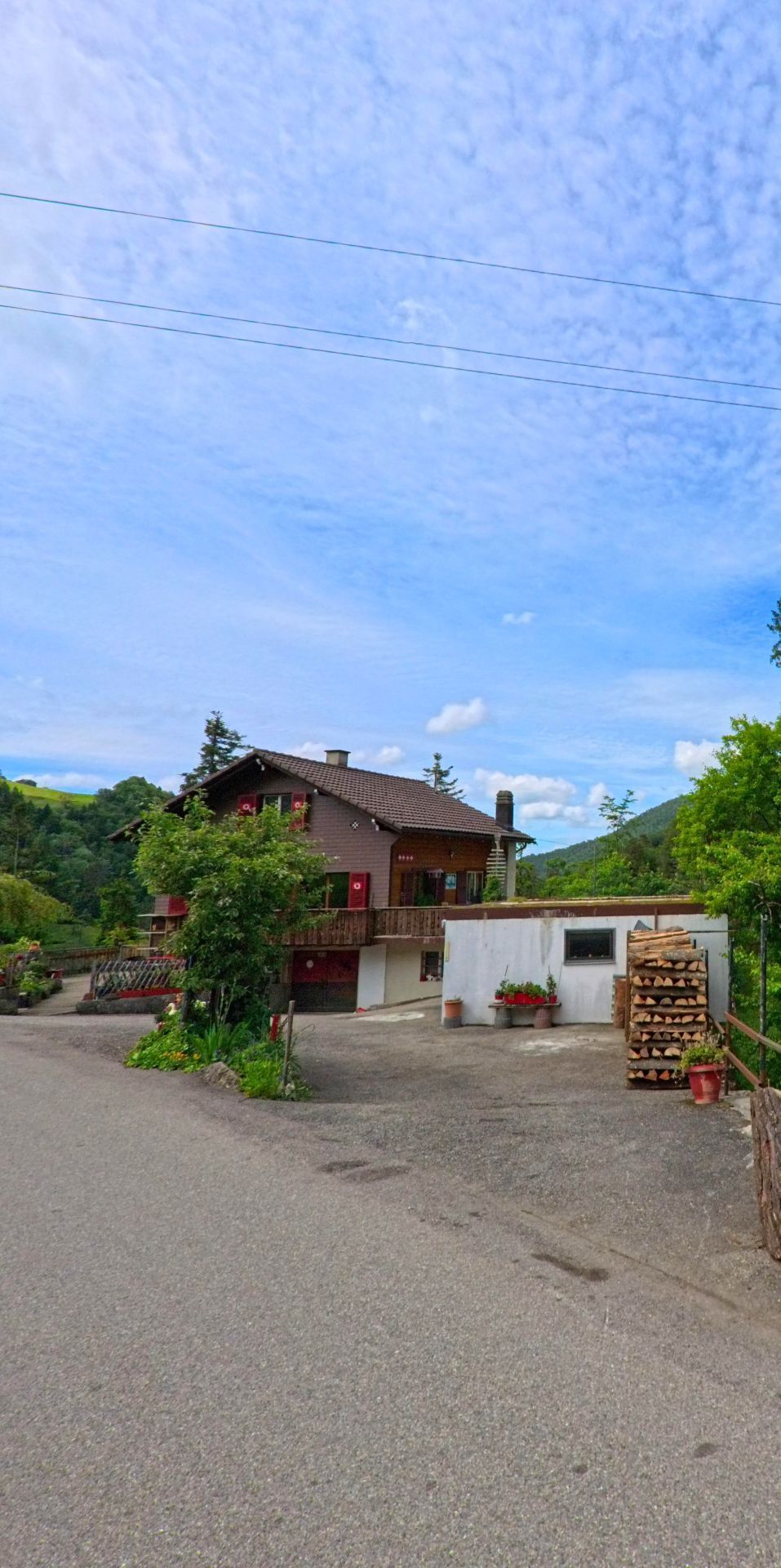 Am Ortsrand von Corcelles (2747, Kanton Bern) steht ein braunes Chalet mit roten Fensterläden, umgeben von Bäumen und einem Vorplatz, auf dem gestapeltes Brennholz und Blumentöpfe angeordnet sind.