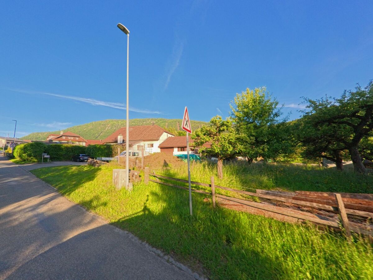 In Corcelles (2747, Kanton Bern) verläuft eine Strasse entlang von Wiesen, Obstbäumen und Wohnhäusern, während ein Verkehrsschild vor Kindern warnt und die Hügel im Hintergrund sichtbar sind.