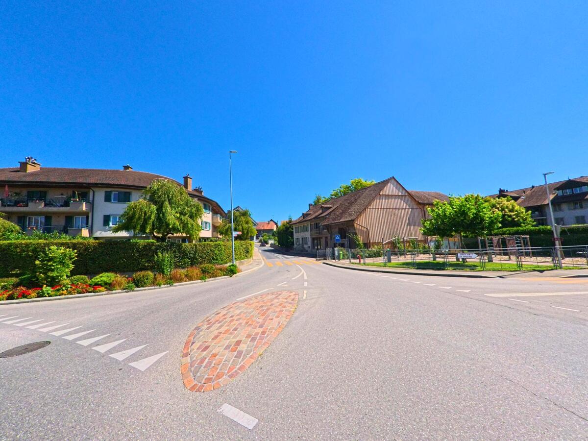 An einer leicht ansteigenden Strasse in Corcelles-pres-Payerne (1562, Kanton Waadt) stehen Mehrfamilienhaeuser und eine grosse Scheune unter blauem Himmel.