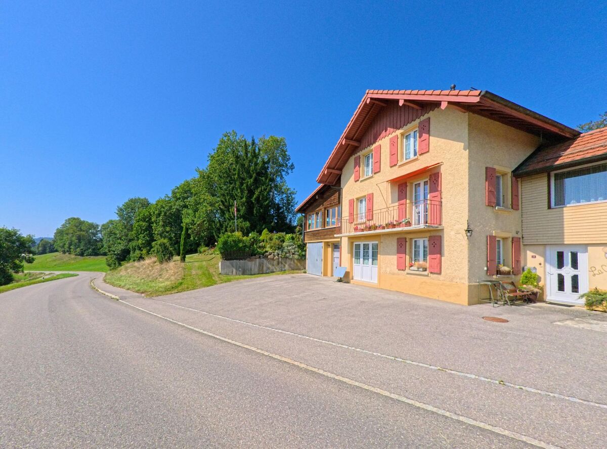 Ein Haus mit roten Fensterlaeden und Balkon steht in Corcelles-le-Jorat (1088, Kanton Waadt) direkt an einer leicht geschwungenen Landstrasse am Ortsrand.