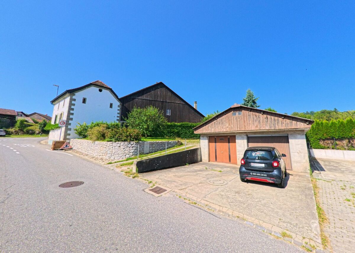 Ein kleines weisses Haus mit Ecksteinen und danebenliegende Scheunenpraegung sind in Corcelles-le-Jorat (1088, Kanton Waadt) an einer Strassenkurve zu sehen, vor einer Garage parkt ein Auto.