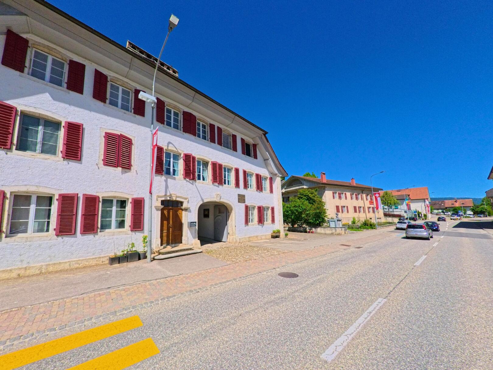 In Corgémont (2606, Kanton Bern) steht ein Gebäude mit roten Fensterläden und historischem Eingang an einer Hauptstrasse mit Kopfsteinpflaster und Zebrastreifen.