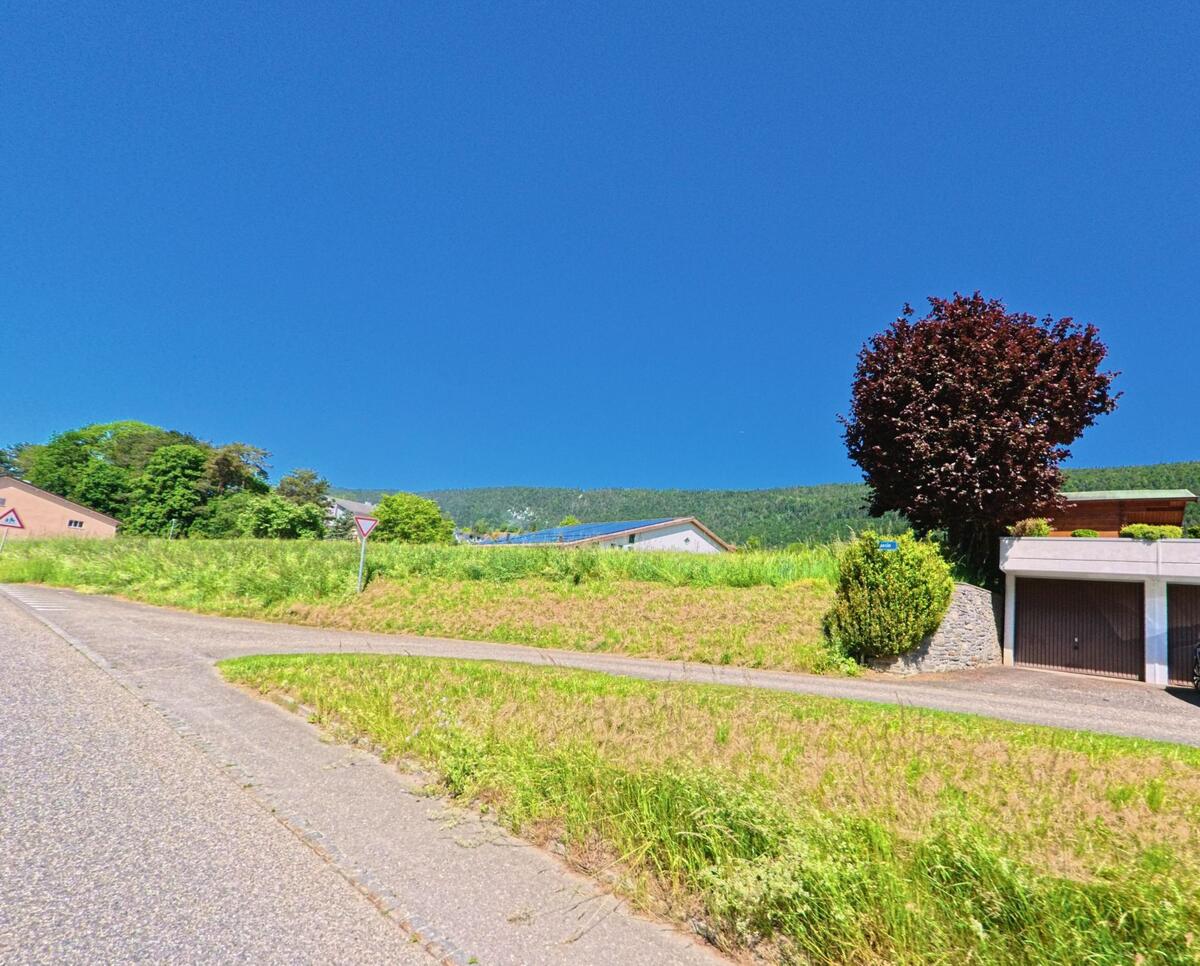 In Corgémont (PLZ 2606, Kanton Bern) steigt ein Seitenweg leicht an, vorbei an Wiesen, Garagen und einem markanten Baum mit rotem Laub.