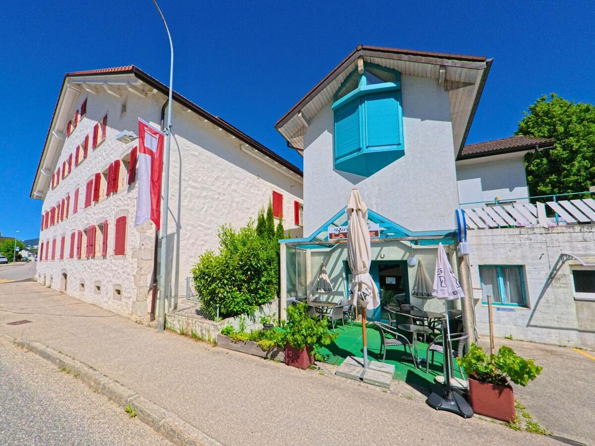 In Corgémont (2606, Kanton Bern) liegt ein kleines Café mit Terrasse unterhalb eines Hauses mit türkisfarbenen Fensterläden, direkt an der Strasse.
