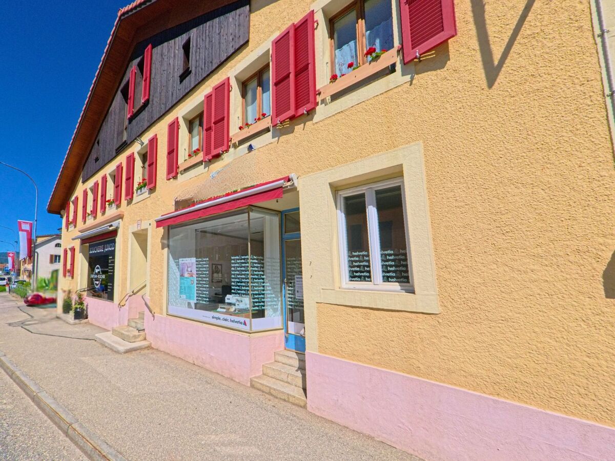 In Corgémont (PLZ 2606, Kanton Bern) beherbergt ein gelb-rosafarbenes Gebäude mit roten Fensterläden mehrere kleine Geschäfte an der Hauptstrasse.