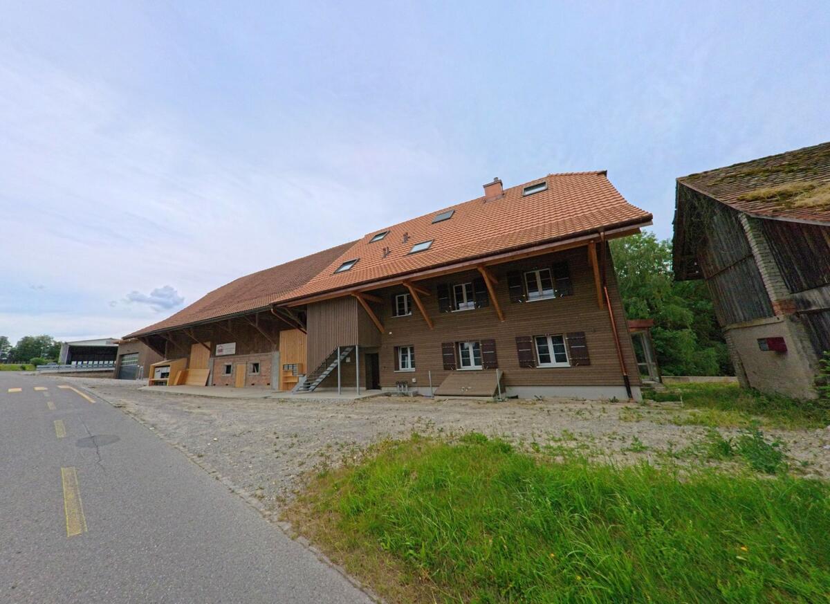 In Corminboeuf (Kanton Freiburg, PLZ 1720) steht ein langgezogenes Bauernhaus mit neuem Ziegeldach und Holzfassade direkt an der Strasse in ländlicher Umgebung.