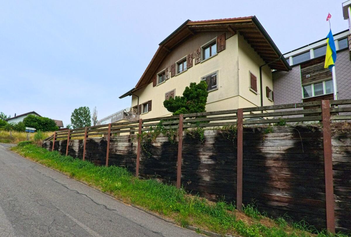 In Corminboeuf (PLZ 1720, Kanton Freiburg) steht ein traditionelles Wohnhaus mit Fensterläden erhöht über einer Stützmauer aus dunklem Holz an einer Nebenstrasse.