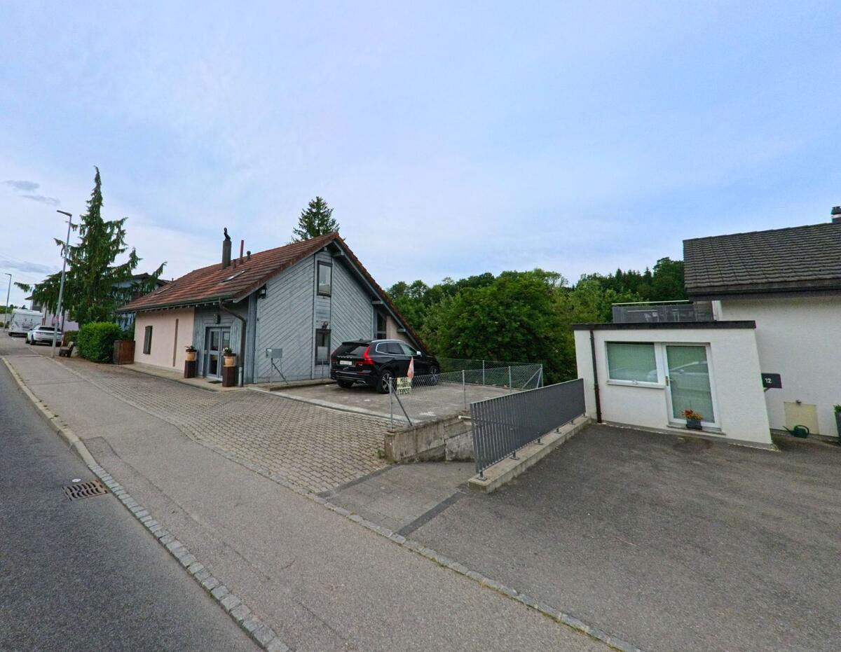 In Corminboeuf (1720, Kanton Freiburg) steht ein kleines Wohnhaus mit Schrägdach und Holzverkleidung an einer Quartierstrasse mit angrenzendem Parkplatz.