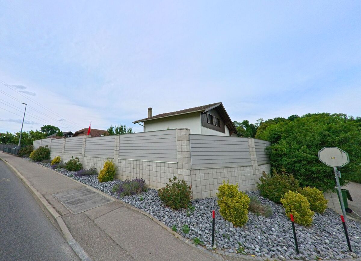 In Corminboeuf (PLZ 1720, Kanton Freiburg) steht ein Einfamilienhaus hinter einer hohen Sichtschutzmauer aus Beton, die entlang des Gehwegs mit Ziersträuchern bepflanzt ist.