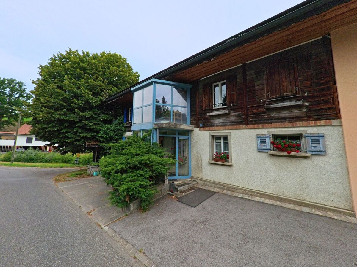 In Corminboeuf (1720, Kanton Freiburg) befindet sich ein traditionelles Wohnhaus mit Holzfassade und einem verglasten Eingangsvorbau an einer ruhigen Seitenstrasse.