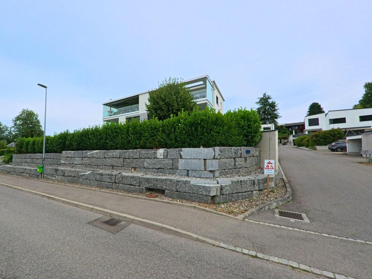 In Corminboeuf (1720, Kanton Freiburg) erhebt sich ein modernes Wohnhaus hinter einer gestuften Natursteinmauer mit gepflegter Hecke entlang einer Nebenstrasse.