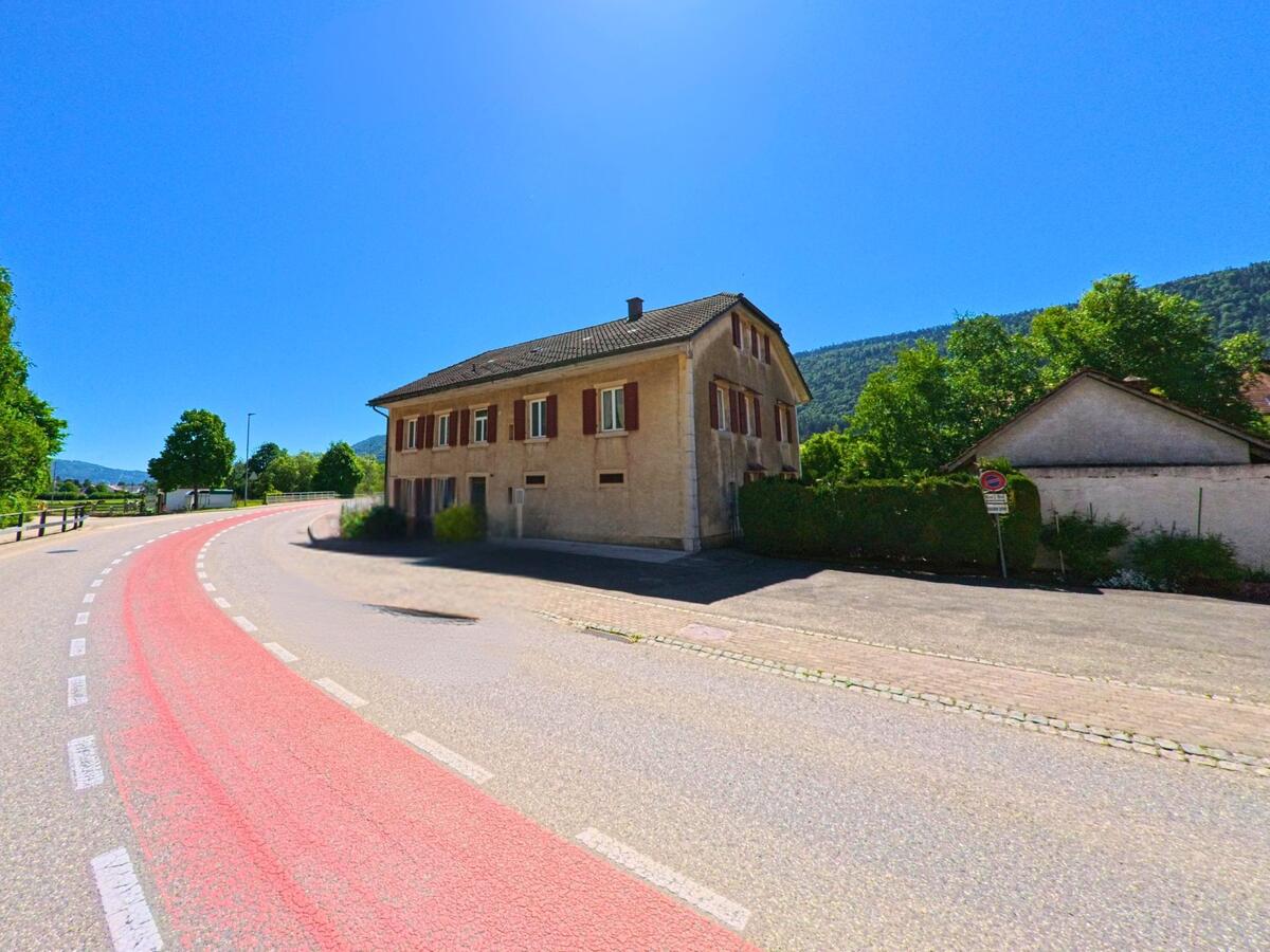 Eine sanft geschwungene Hauptstrasse führt in Cormoret (2612, Kanton Bern) an einem schlichten Wohnhaus mit roten Fensterläden vorbei.