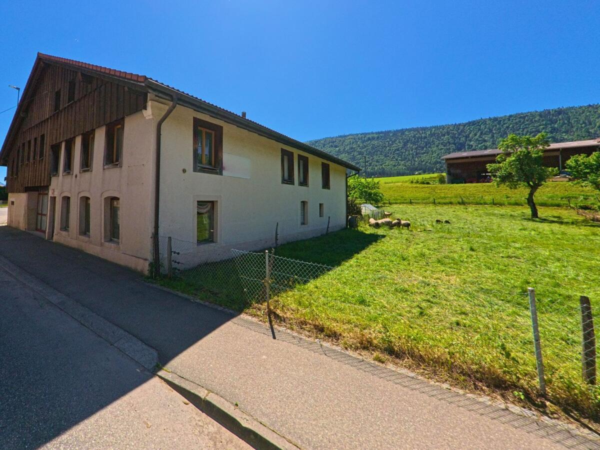 In Cormoret (2612, Kanton Bern) steht ein Wohnhaus mit angrenzender Weide, auf der einige Schafe unter blauem Himmel grasen.