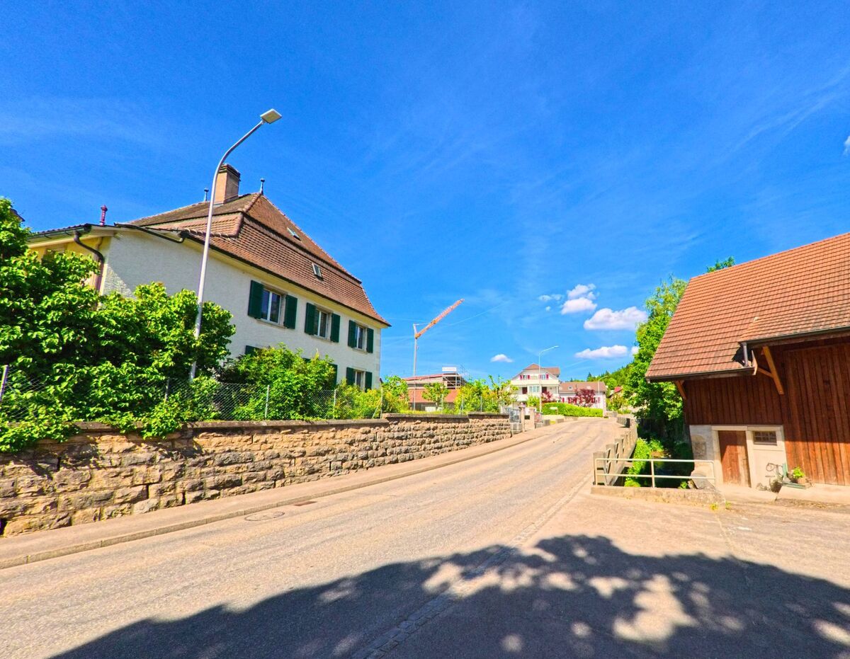 In Cornol (2952, Kanton Jura) führt eine Straße an einem historischen Gebäude und einem modernen Bauprojekt vorbei, während ein Stück Land links von einem Zaun begrenzt wird.