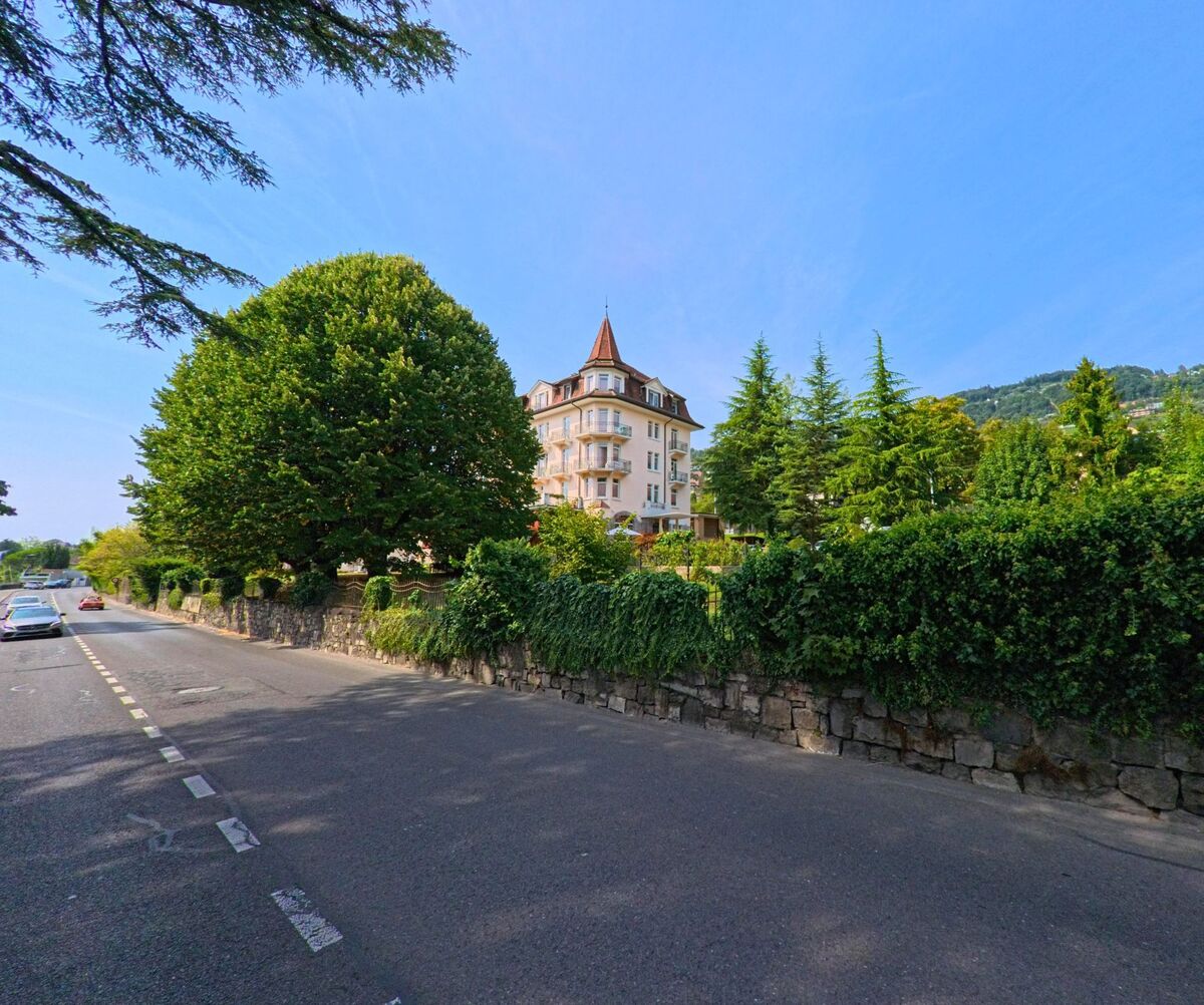 Ein grosses Gebäude mit Türmchen erhebt sich hinter hohen Bäumen an einer Strasse in Corseaux (1802, Kanton Waadt).