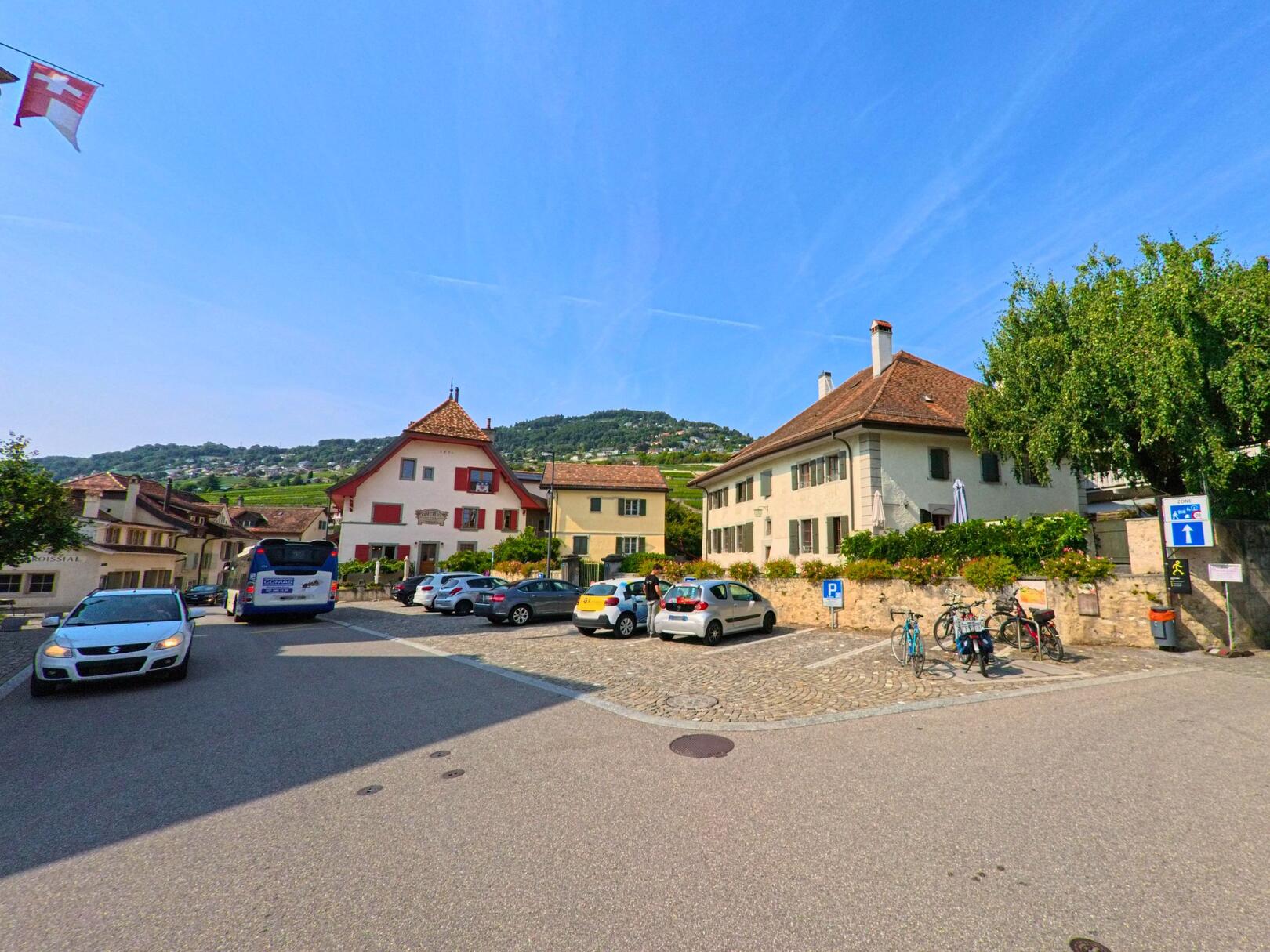 Im Zentrum von Corsier-sur-Vevey (1804, Kanton Waadt) liegt ein Platz mit parkenden Autos und historischen Wohnhaeusern vor dem Hintergrund der Weinberge.