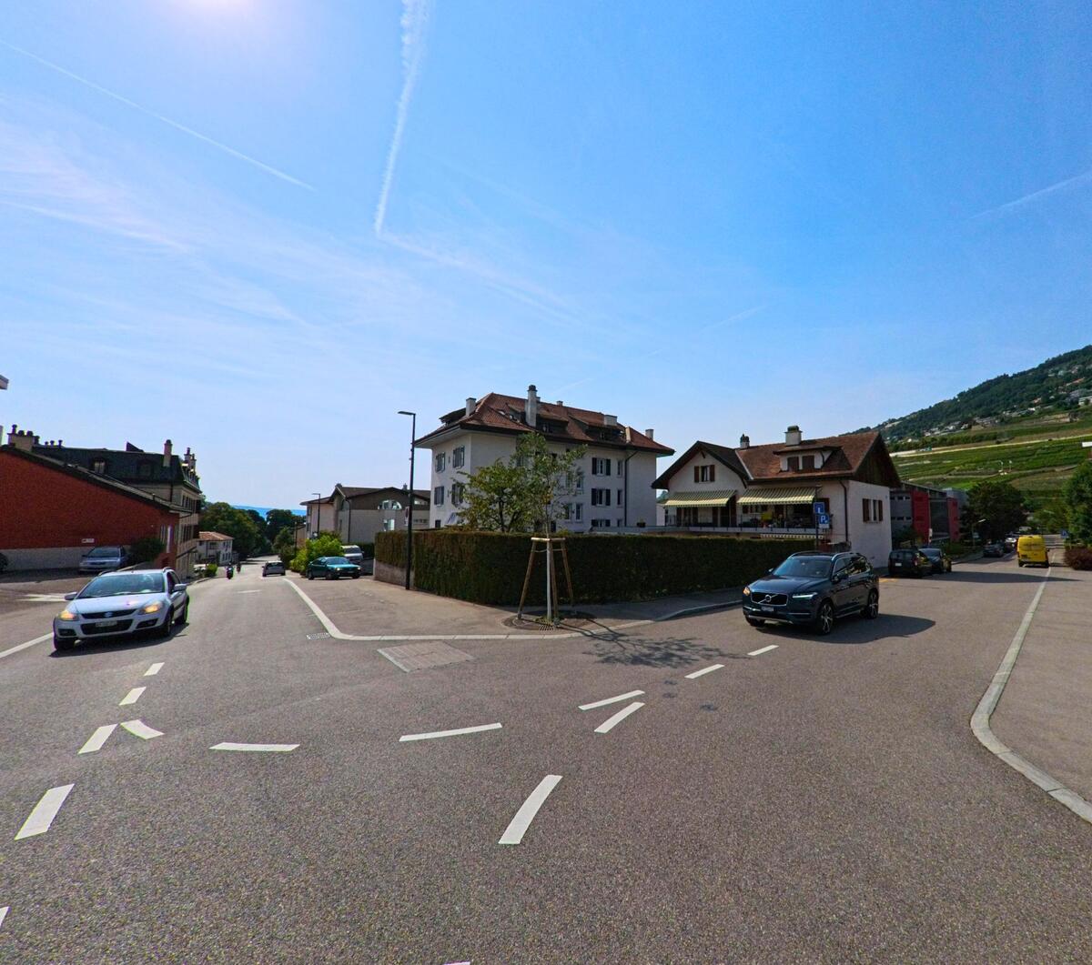 An einer breiten Strassenkreuzung in Corsier-sur-Vevey (1804, Kanton Waadt) stehen Wohnhaeuser, waehrend im Hintergrund Weinberge den Hang saeumen.
