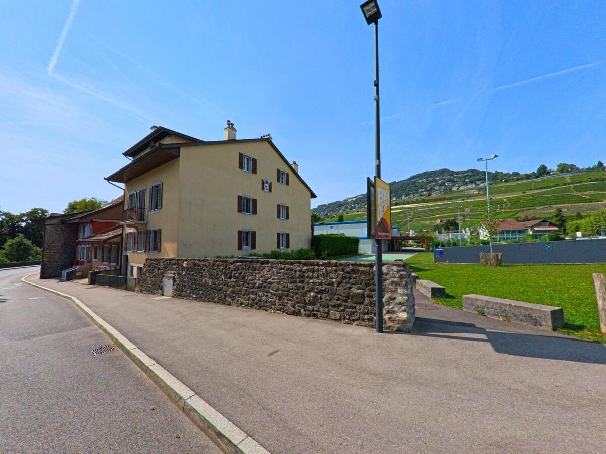Ein gelbes Wohnhaus mit braunen Fensterlaeden steht in Corsier-sur-Vevey (1804, Kanton Waadt) neben einer Strasse, waehrend im Hintergrund Weinberge am Hang zu sehen sind.