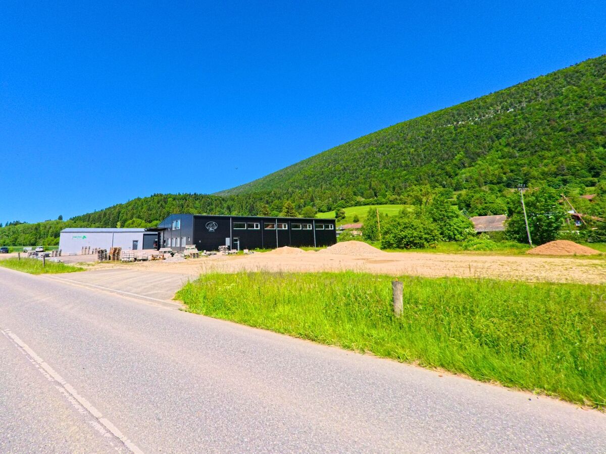 Am Rand von Cortébert (2607, Kanton Bern) liegen moderne Industriehallen vor einem dicht bewaldeten Hang, umgeben von Wiesen und Schotterflächen.