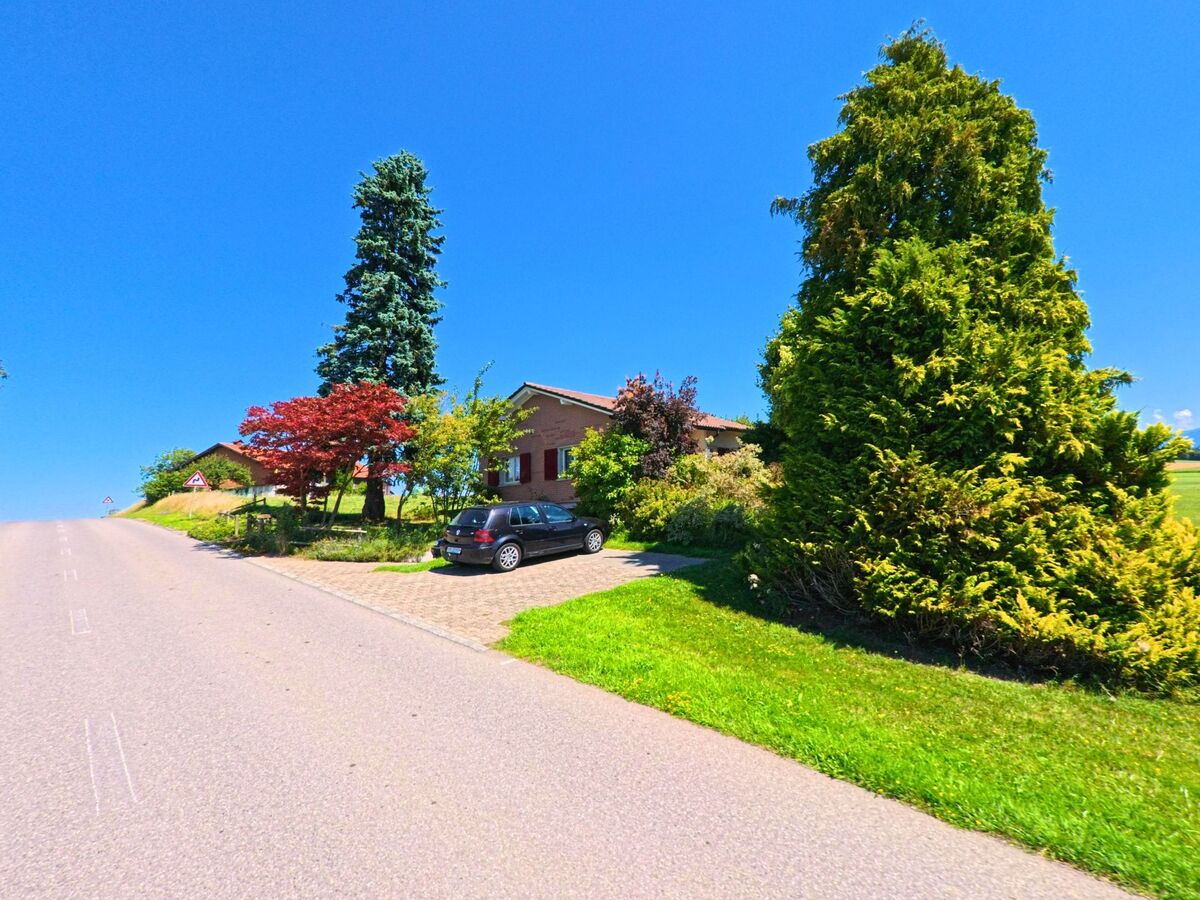 Ein Wohnhaus mit rotem Backstein und gepflegtem Garten liegt an einer ansteigenden Strasse in Cottens (1741, Kanton Freiburg), flankiert von grossen Nadelbaeumen und buntem Laub.