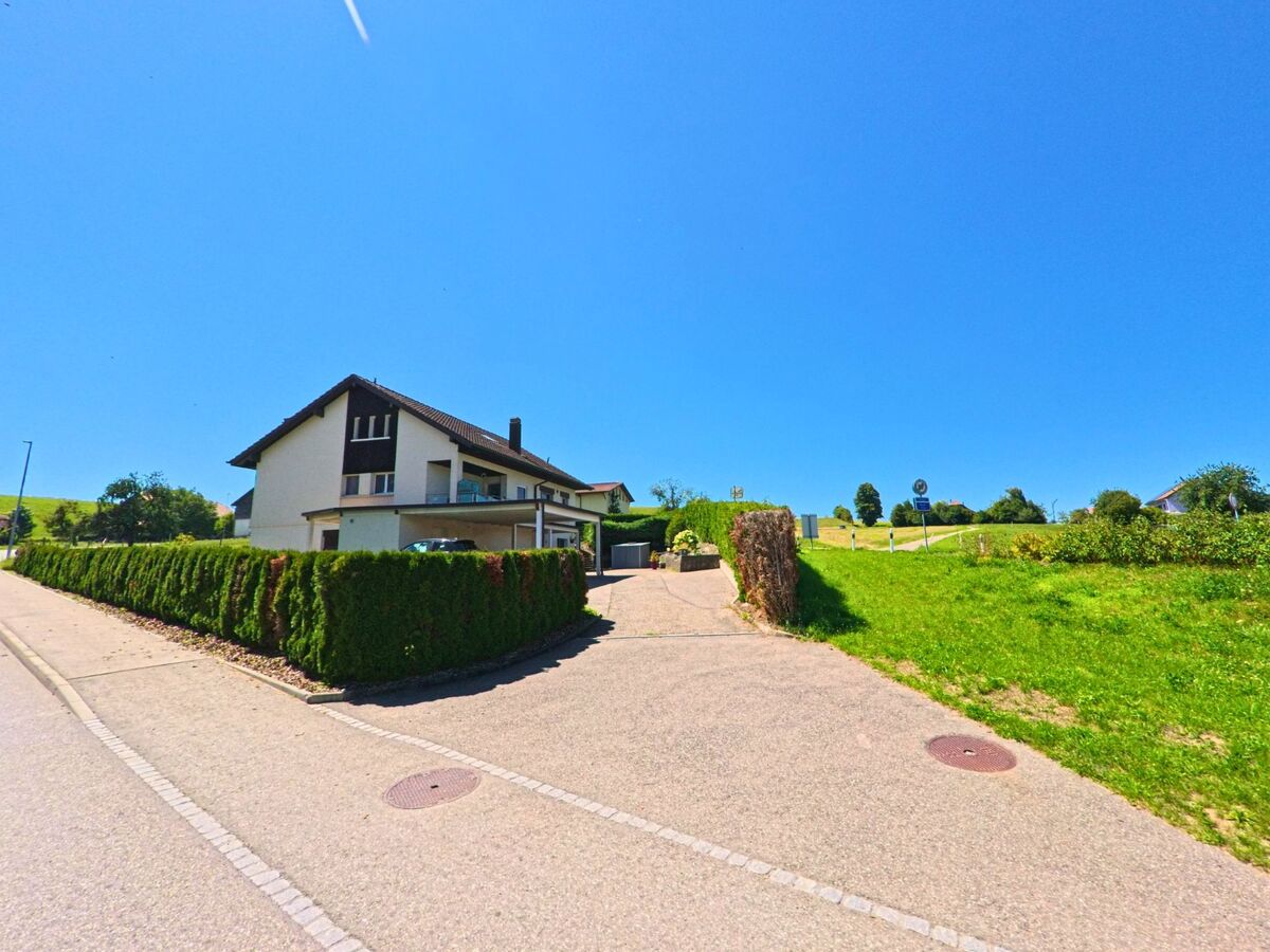Ein modernes Einfamilienhaus mit dunklem Dach und gepflegter Thujahecke steht am Rand einer ruhigen Strasse in Cottens (1741, Kanton Freiburg) neben einer offenen Wiese.