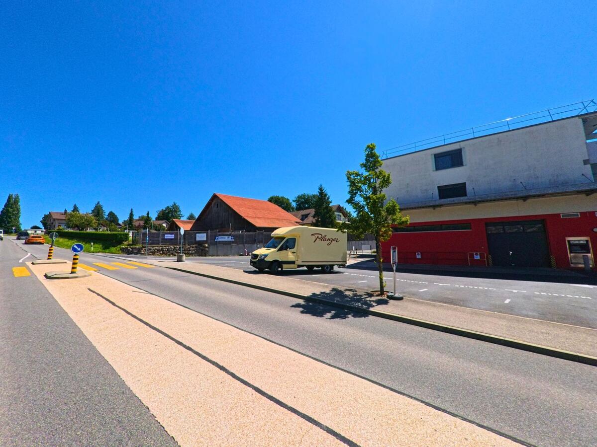 An einer Hauptstrasse in Cottens (1741, Kanton Freiburg) stehen ein rotes Gewerbegebäude und eine Scheune mit rotem Dach gegenüber, neben einer Verkehrsinsel mit Fussgaengerstreifen.