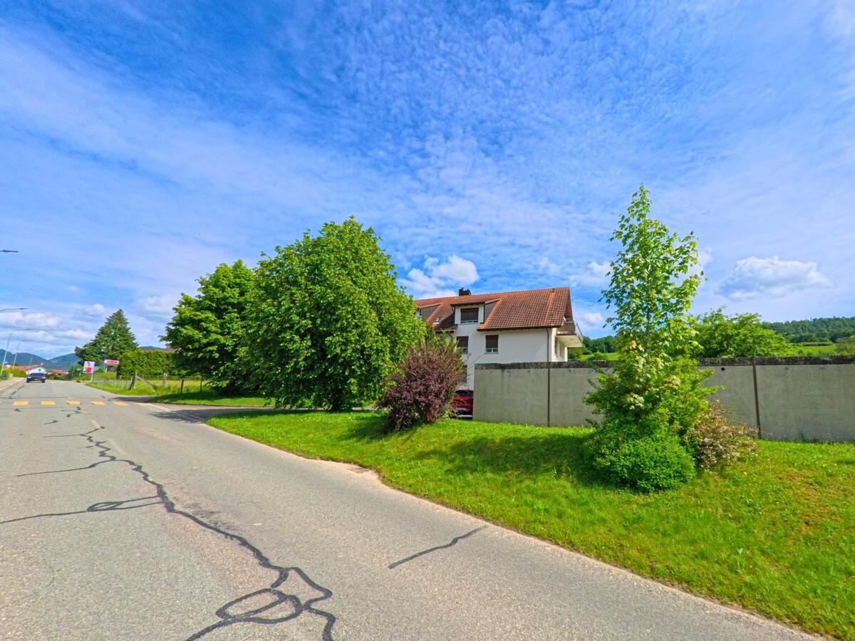 Am Ortseingang von Courchapoix (2825, Kanton Jura) führt eine leicht beschädigte Strasse an einer Lärmschutzwand und einem Wohnhaus mit rotem Ziegeldach vorbei.