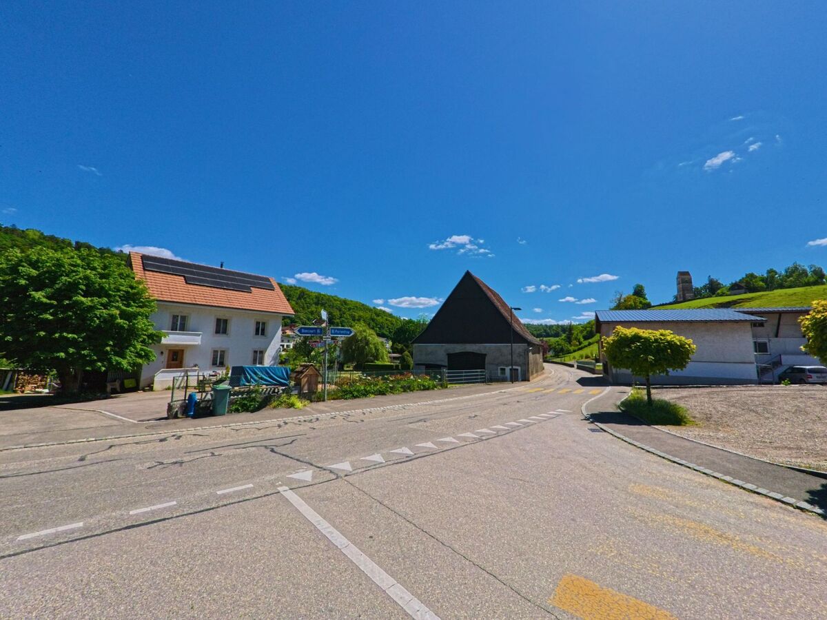 Im Dorfzentrum von Courchavon (2922, Kanton Jura) treffen sich mehrere Strassen bei einem Wohnhaus mit Solarpanels, einer alten Scheune und einem Wegweiser.
