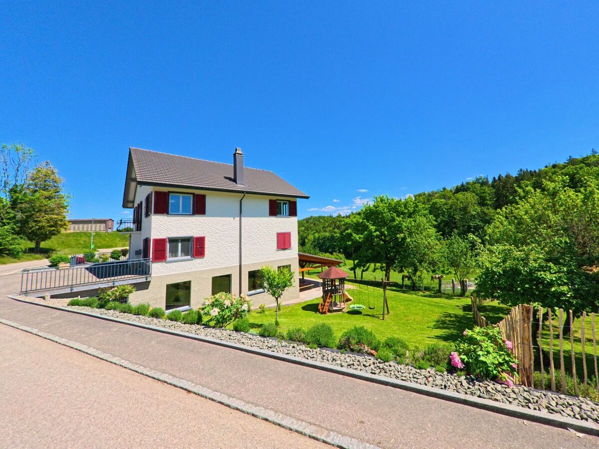 Ein modernes Wohnhaus mit roten Fensterläden steht in Courchavon (2922, Kanton Jura) an einer Hangstrasse, daneben ein gepflegter Garten mit Spielplatz.