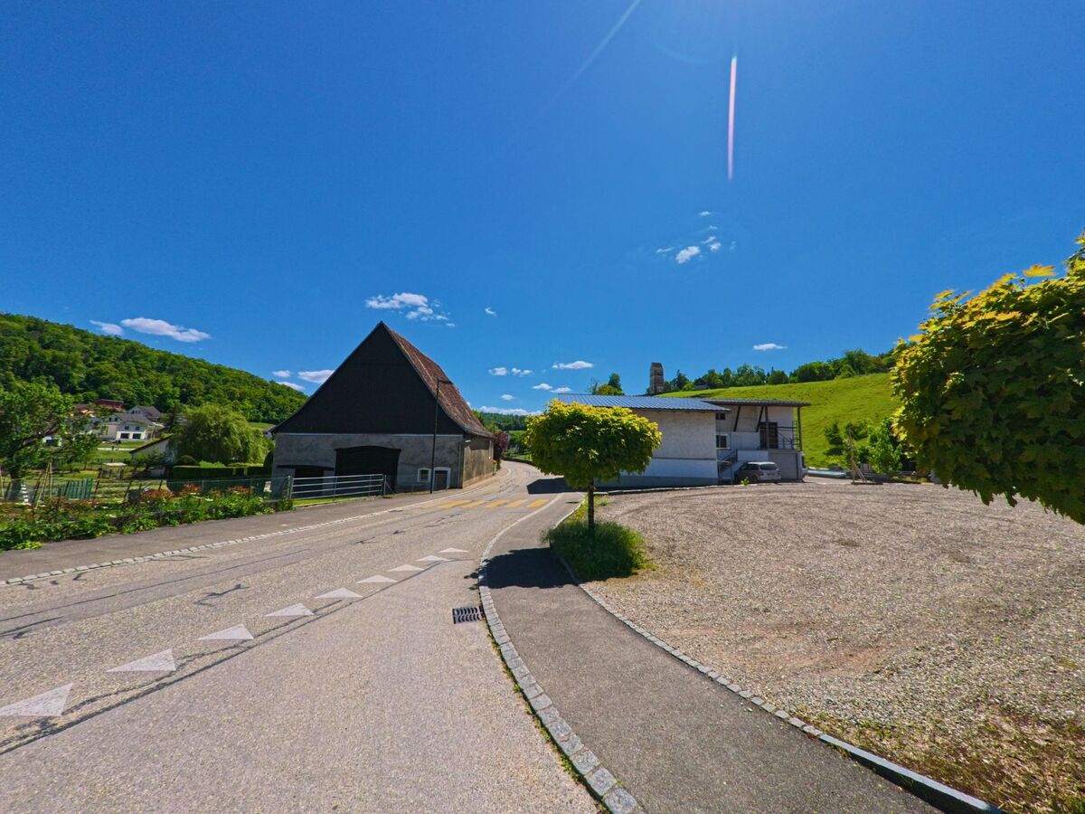 In Courchavon (2922, Kanton Jura) führt eine leicht geschwungene Strasse an einer alten Scheune und einem modernen Haus vorbei durch das grüne Hügelland.