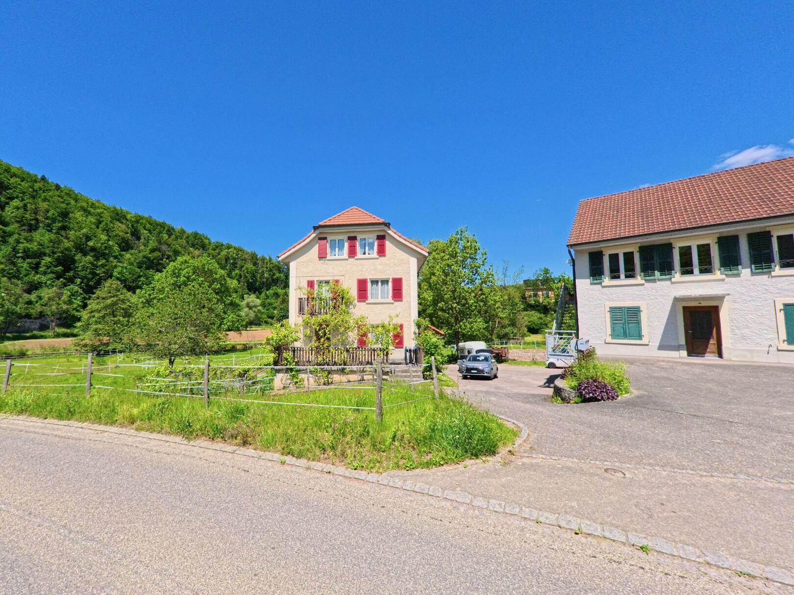 In Courchavon (2922, Kanton Jura) stehen zwei gepflegte Gebäude mit farbigen Fensterläden an einer ruhigen Strasse, umgeben von Wiesen und bewaldeten Hügeln.