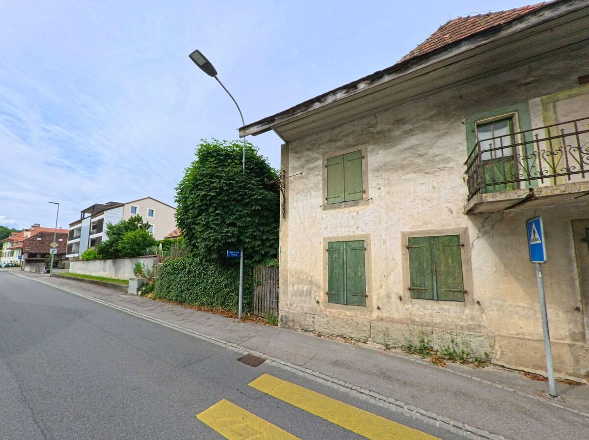An einer Hauptstrasse in Courgevaux (1796, Kanton Freiburg) steht ein älteres Gebäude mit verwitterter Fassade und geschlossenen grünen Fensterläden gegenüber modernen Wohnhäusern.