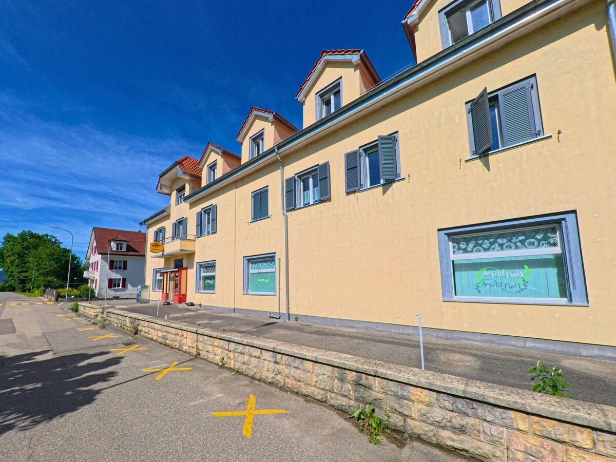 In Courrendlin (2830, Kanton Jura) befindet sich ein gelbes Gebaeude mit Fensterlaeden und Ladeneingang entlang einer Strasse mit markierten Parkflaechen.