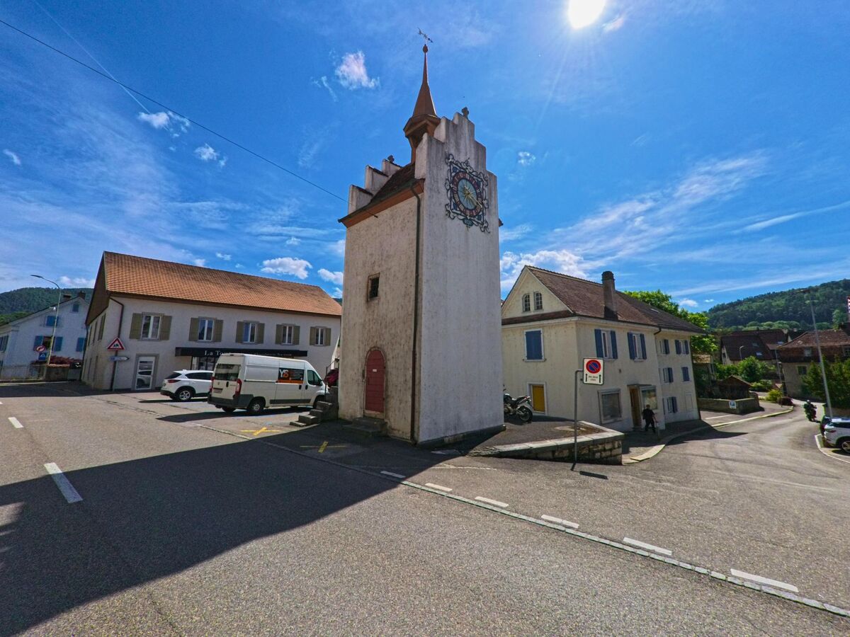 Im Zentrum von Courrendlin (2830, Kanton Jura) steht ein historischer weisser Turm mit bemalter Uhr, umgeben von Wohnhaeusern und kleinen Geschaeften.
