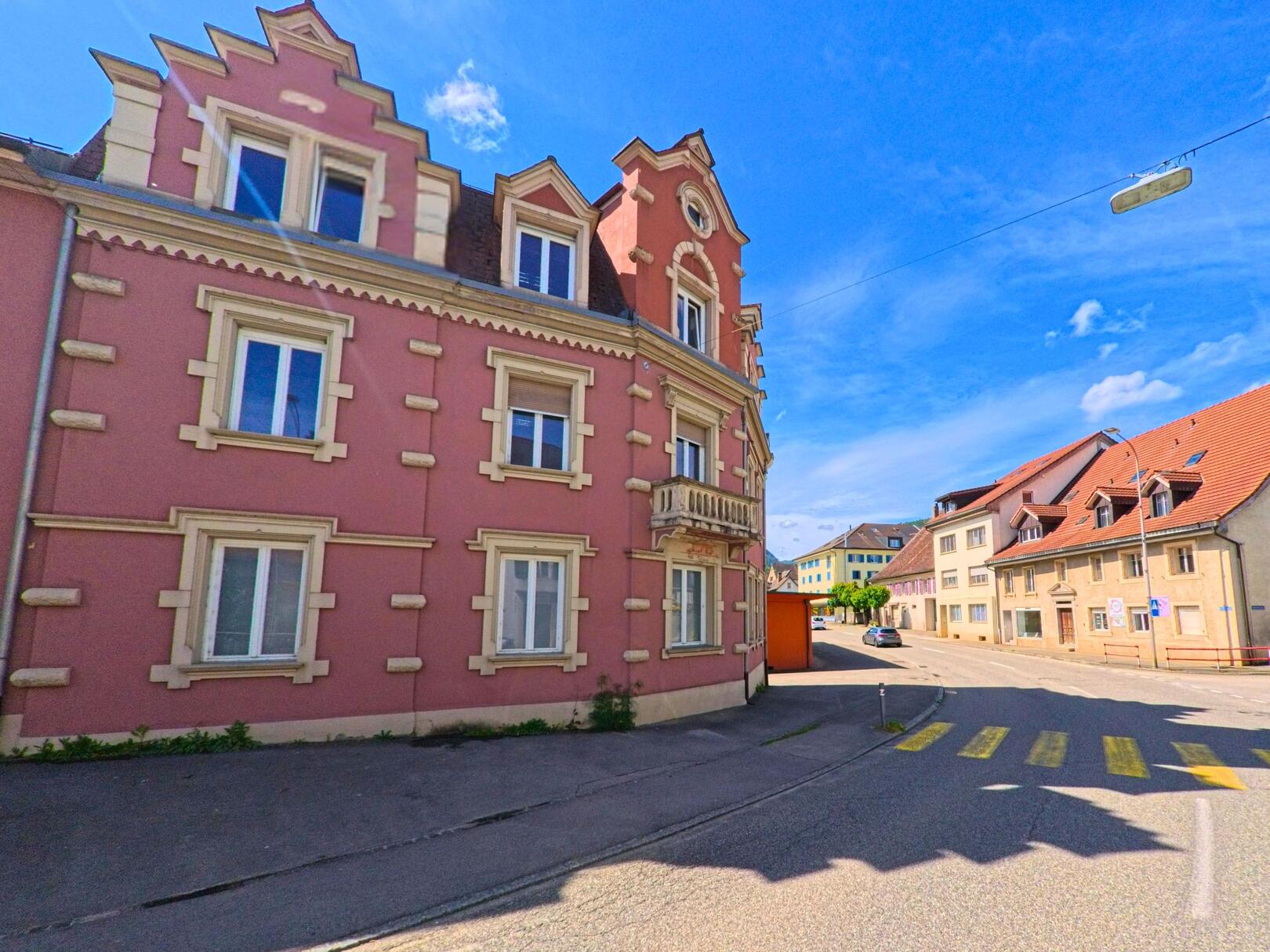 In Courrendlin (2830, Kanton Jura) stehen entlang einer leicht geschwungenen Strasse mehrere gut erhaltene Wohnhaeuser, darunter ein auffaellig rosafarbenes Gebaeude mit dekorativer Fassade.