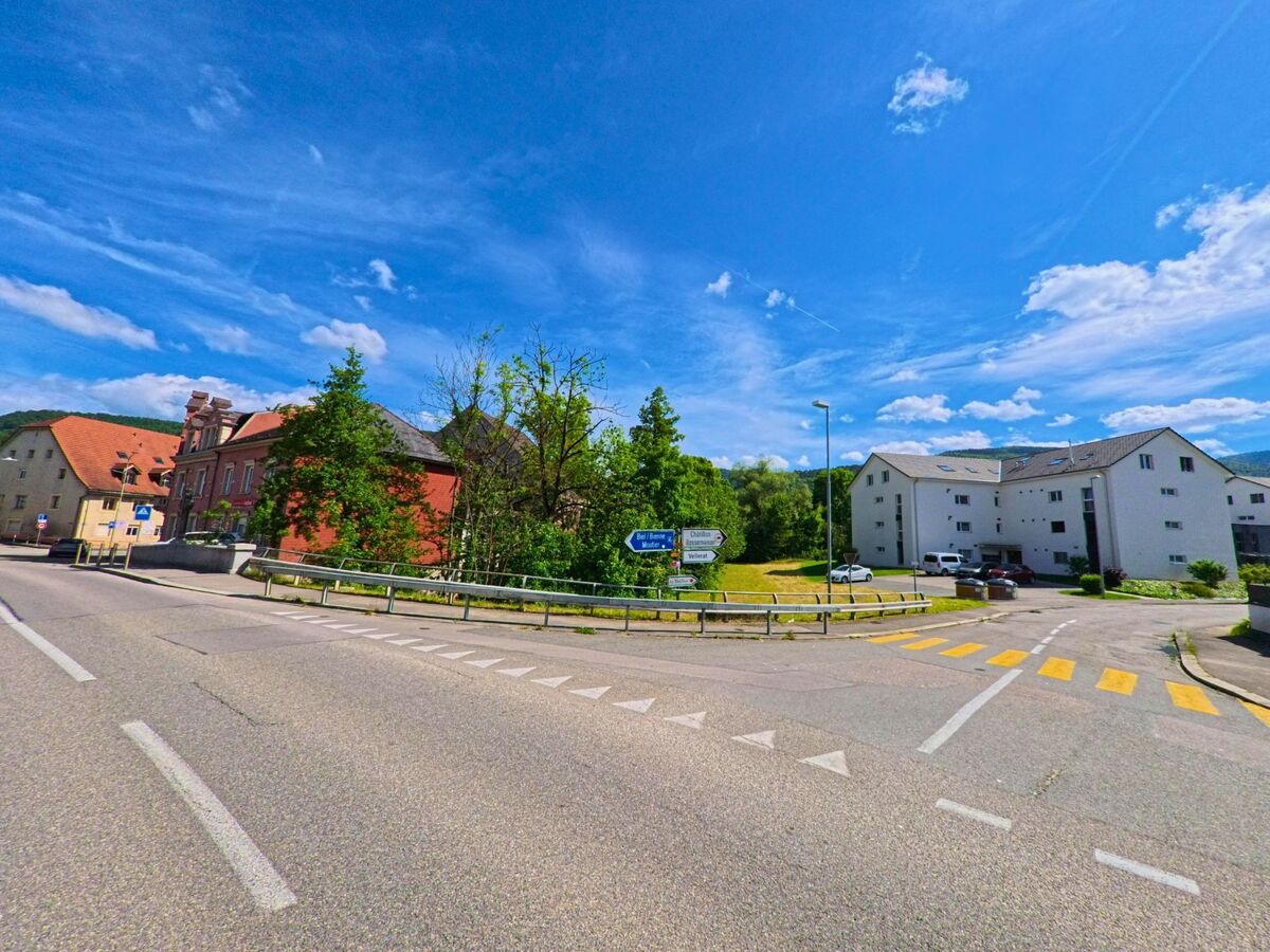 In Courrendlin (2830, Kanton Jura) trifft eine Einmündung mit Fussgaengerstreifen auf eine Hauptstrasse, flankiert von historischen und modernen Gebaeuden sowie Wegweisern nach Delémont und Vellerat.