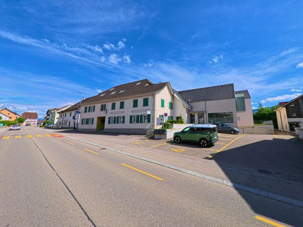 In Courroux (2822, Kanton Jura) zeigt sich ein zentral gelegenes Gebaeudeensemble mit Parkplaetzen und einem Zebrastreifen bei klarem Himmel.